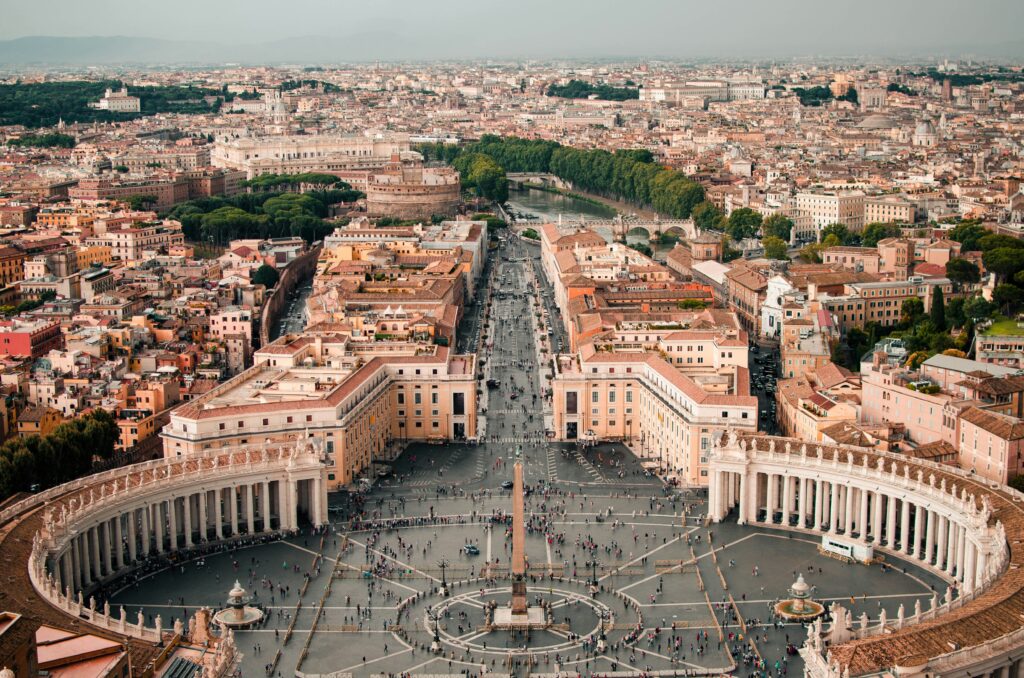 High-angle aerial panoramic view of St. Peter's Square (Piazza San Pietro) in Vatican City, showing the elliptical colonnades, central obelisk, crowds of visitors, and the Via della Conciliazione leading into Rome under clear skies.