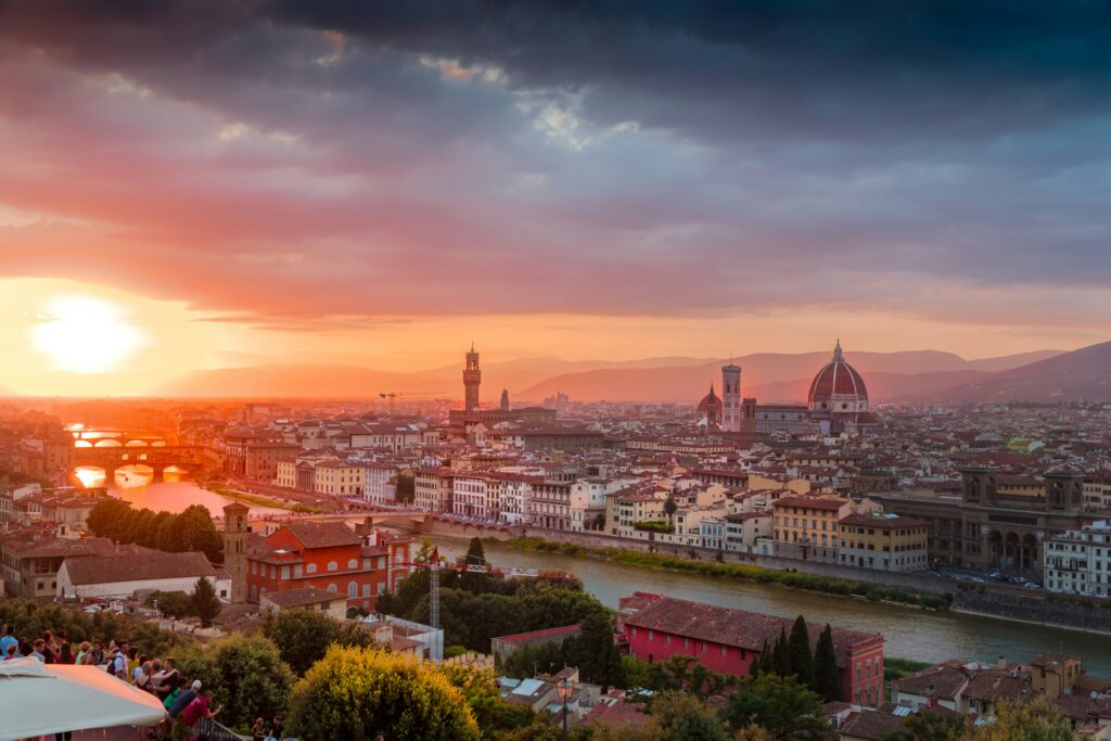 A sweeping, elevated, and unpopulated panoramic view of the Florence skyline at a serene sunset. The iconic Duomo and Giotto's Bell Tower, Palazzo Vecchio, and Ponte Vecchio along the River Arno are clearly visible under a dramatic, cloud-filled sky with rich orange and pink tones. The foreground viewing terrace, previously with people and an umbrella, is pristine and secluded. All crowds and modern clutter have been removed to present a clean, undisturbed, and deeply romantic cityscape perfect for an exclusive luxury couple's getaway.