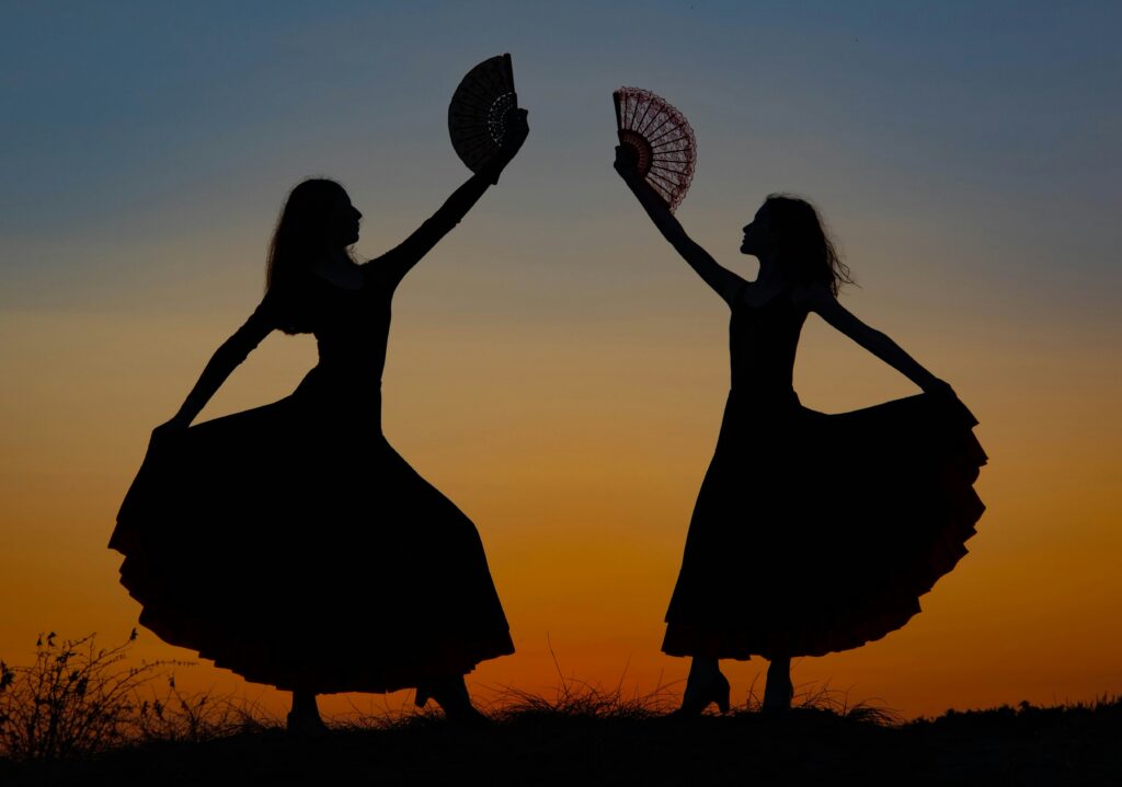 Silhouette of two female dancers in traditional long dresses holding fans high against a dramatic orange and blue sunset, symbolizing an authentic Seville Flamenco experience for couples.