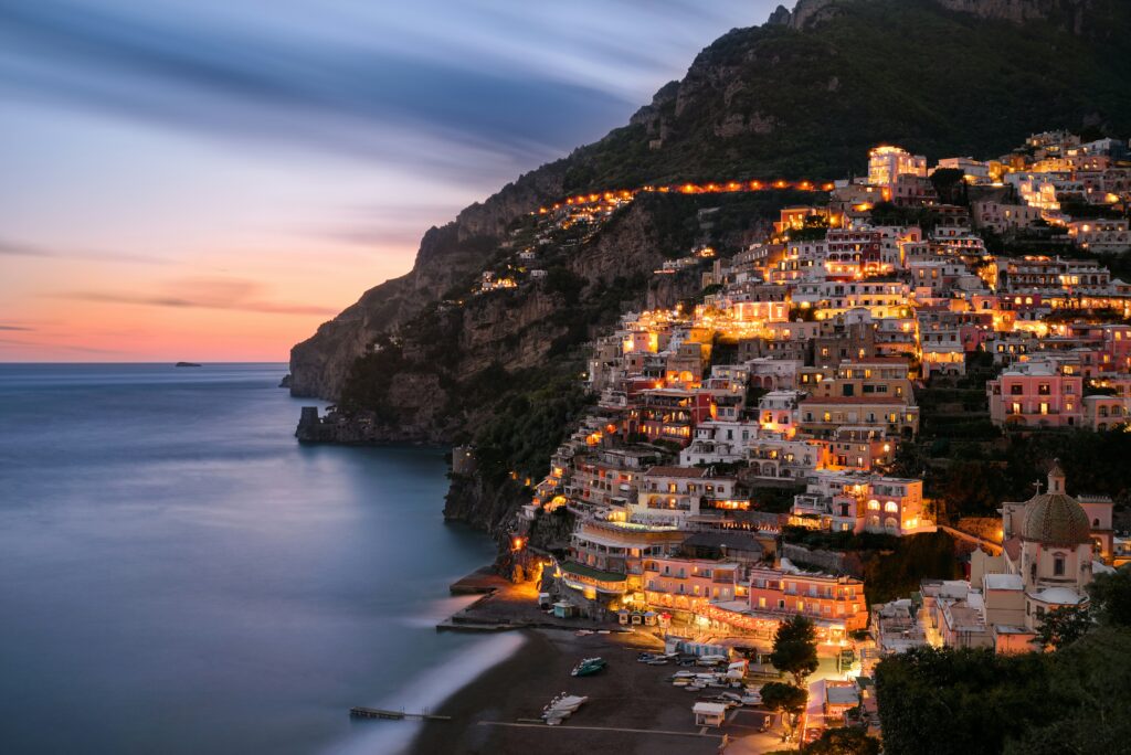 Panoramic sunset view of Positano on the Amalfi Coast, Italy, featuring illuminated luxury hotels and colorful houses cascading down the cliff to the sea, representing one of the best romantic coastal towns for couples.