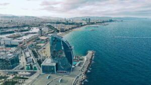 An aerial high-angle panoramic view of the Barcelona coastline in Spain, highlighting the iconic sail-shaped glass architecture of the W Barcelona hotel standing next to the blue Mediterranean Sea and the sandy beaches stretching into the city skyline under a cloudy sky.