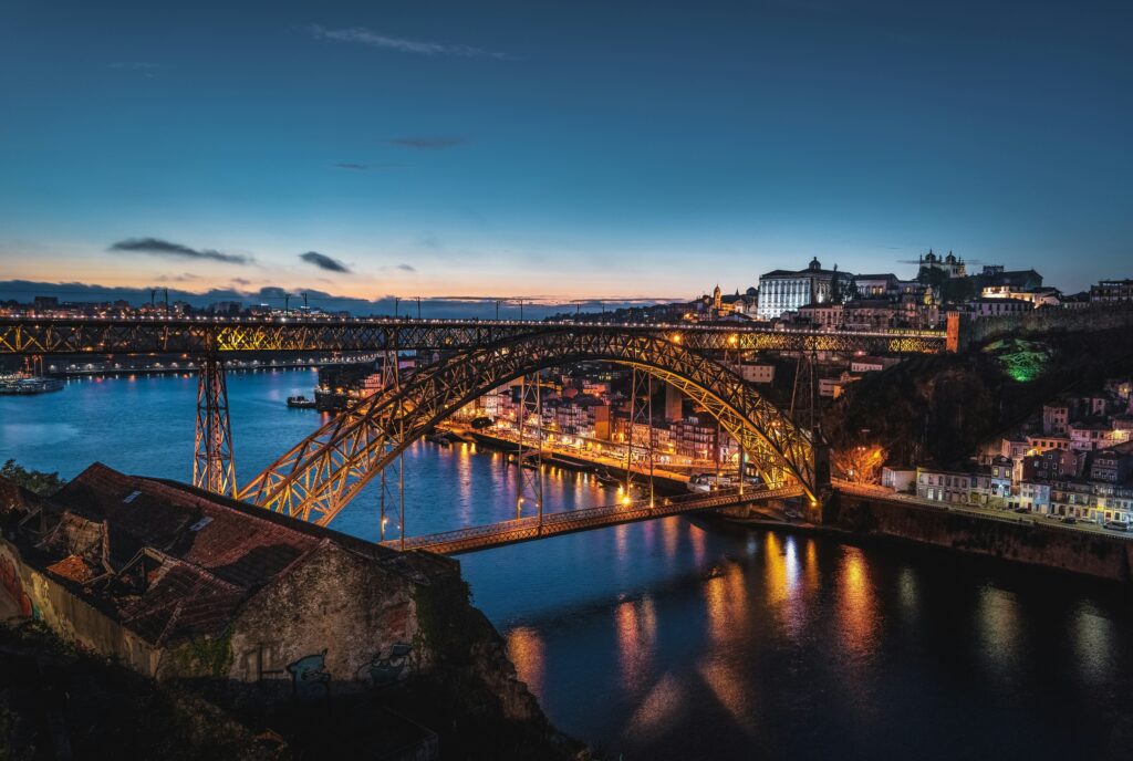 Panoramic twilight view of Porto and the illuminated Dom Luís I Bridge over the Douro River. The golden lights of the historic Ribeira district and the city skyline reflect on the calm dark blue water under a sunset sky.
