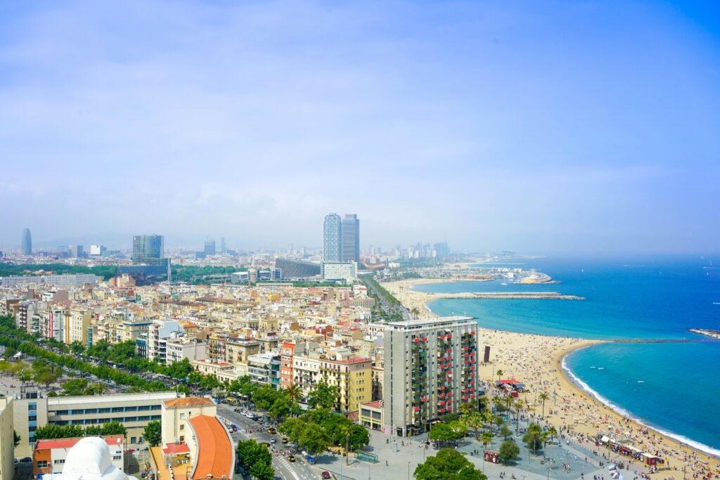High-angle panoramic view of Barcelona's coastline, featuring a crowded sandy beach meeting the vibrant blue Mediterranean Sea, coastal city buildings, and a bright clear sky.
