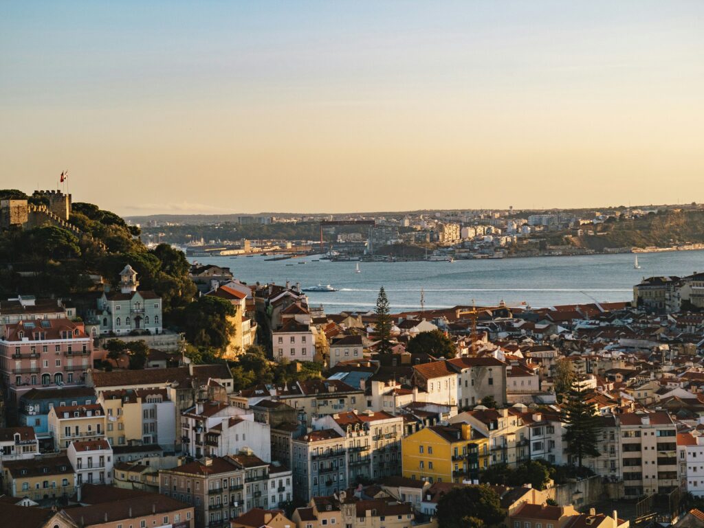 Panoramic view of Lisbon hills and traditional colorful buildings during a golden hour sunset. Castelo de São Jorge sits on the left hill overlooking the historic Alfama district, while the Tagus River with a sailing boat stretches across the background.