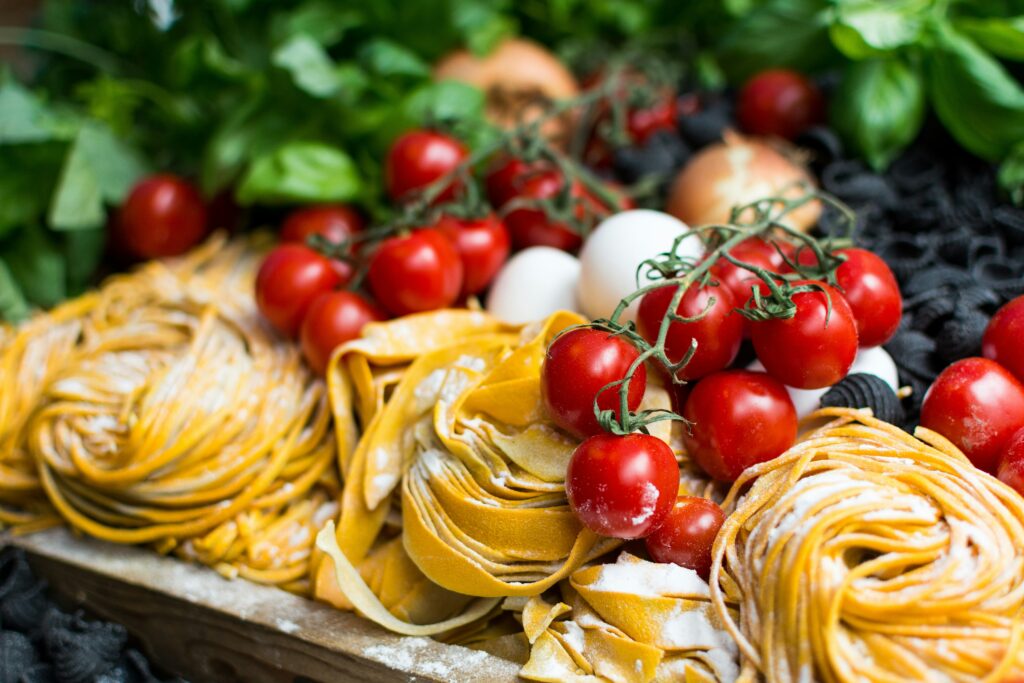 Close-up shot of fresh, uncooked artisanal Italian pasta nests dusted with flour, surrounded by bright red cherry tomatoes on the vine, white eggs, and fresh green basil leaves.