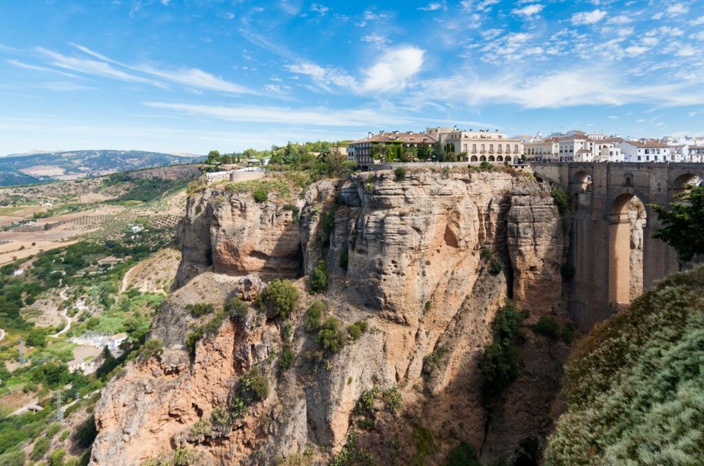 Panoramic view of the historic town of Ronda, Spain, perched dramatically on top of massive cliffs. The image shows white buildings, the famous Puente Nuevo stone bridge spanning the deep gorge, and a lush green valley extending into the horizon under a blue sky.
