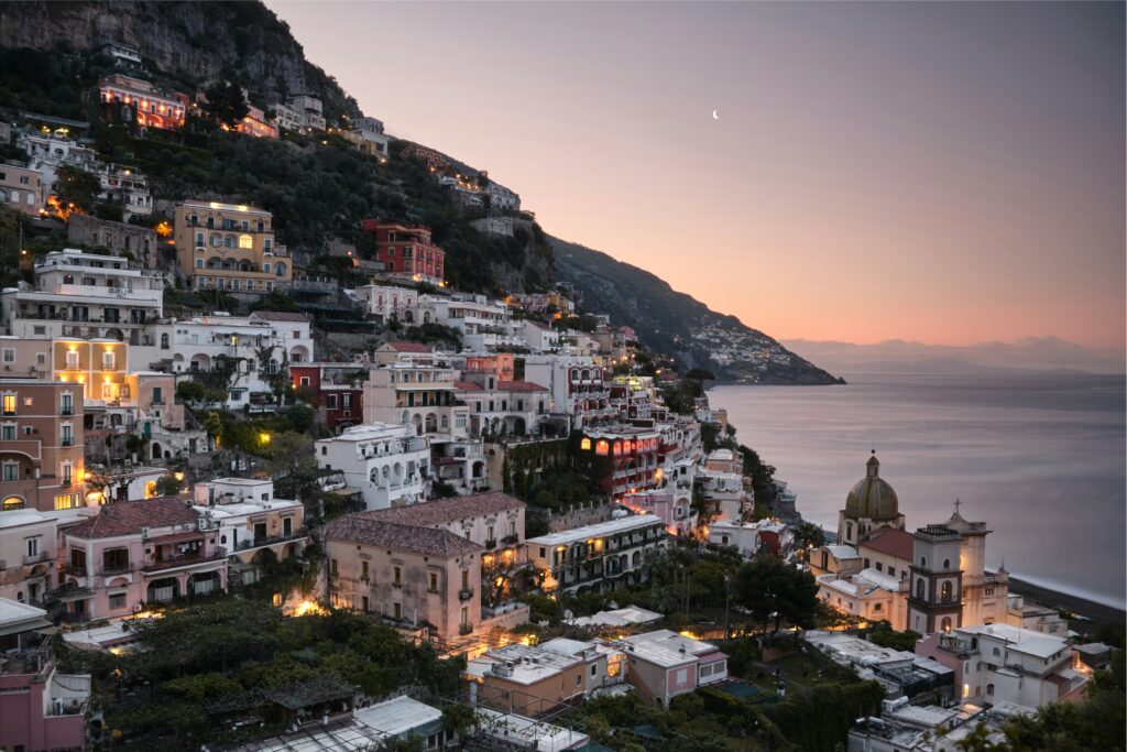 Panoramic twilight view of Positano on the Amalfi Coast, Italy, showcasing luxury cliffside hotels and pastel-colored houses illuminated with warm lights next to a calm sea under a soft evening sky.