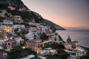Panoramic twilight view of Positano on the Amalfi Coast, Italy, showcasing luxury cliffside hotels and pastel-colored houses illuminated with warm lights next to a calm sea under a soft evening sky.