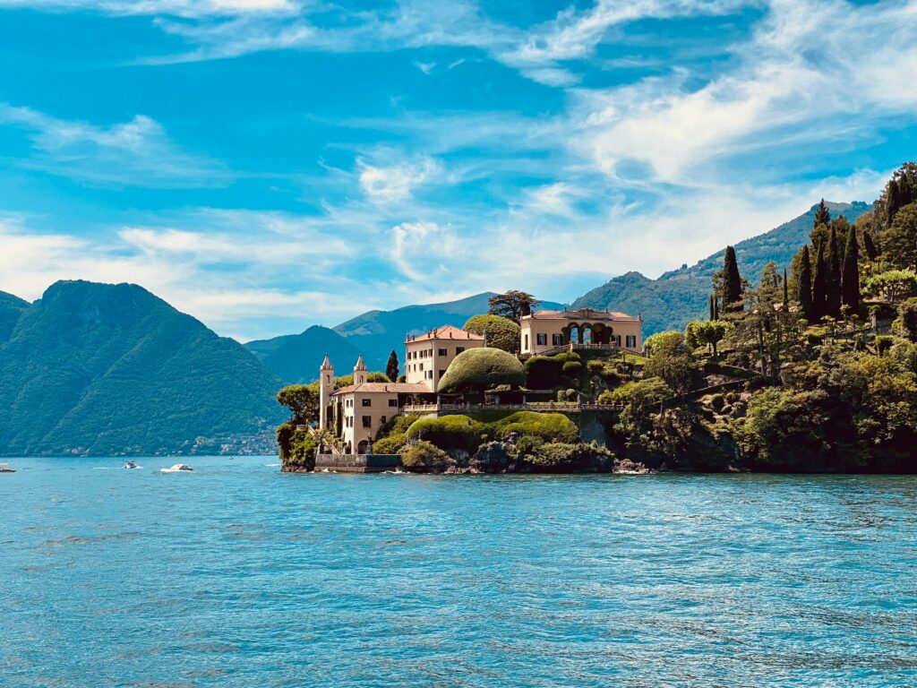 A breathtaking lakeside view of the historic Villa Balbianello on Lake Como, Italy, showcasing terraced gardens, elegant architecture, and lush green trees against a bright blue sky with wispy clouds.