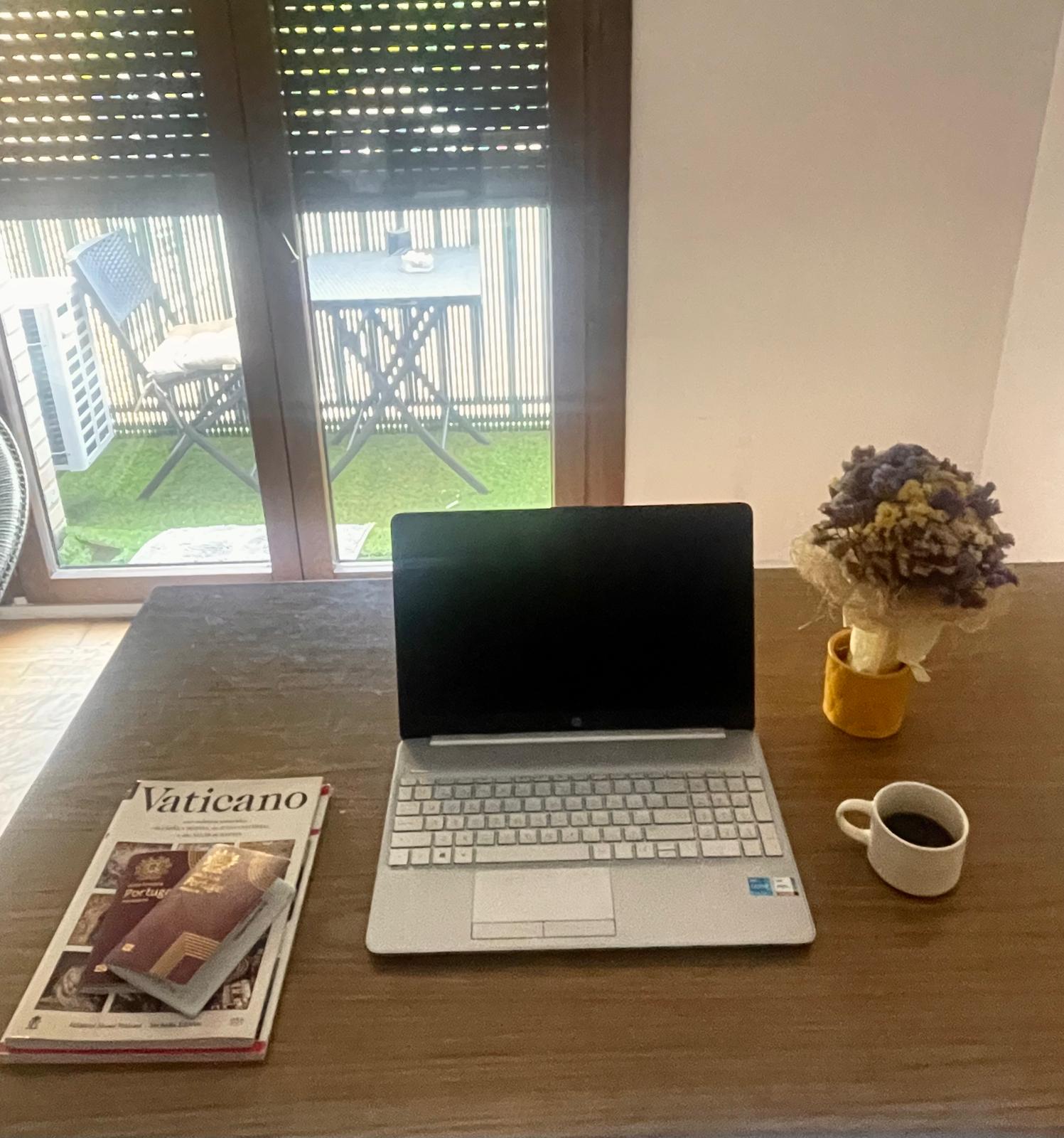 A wood desk with a laptop, a coffee, two books unde twos passports with a window in the background.
