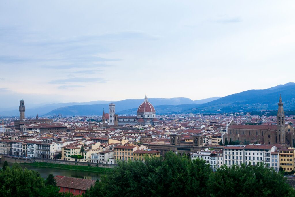 Panoramic high-angle view of the Florence skyline in Italy, featuring the iconic red dome of the Santa Maria del Fiore Cathedral (Duomo), Giotto's Campanile, and the Palazzo Vecchio tower rising above historic terracotta rooftops and the Arno River under a soft sky.