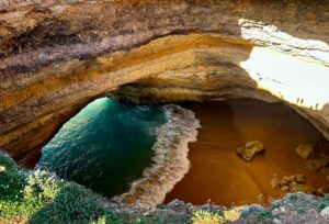 High-angle looking down into the dramatic Benagil Sea Cave in the Algarve, showing the sunlit golden rock arches, a small secluded sandy beach, and ocean waves crashing inside with vivid turquoise water.