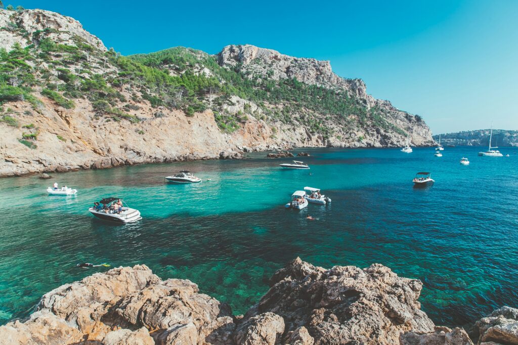 An elevated landscape photograph of a stunning turquoise and blue Mediterranean bay in Mallorca, Spain, surrounded by high, steep rocky cliffs dotted with green pine trees. Several leisure boats and yachts are anchored in the crystal-clear water under a bright blue sky, with rocky foreground elements framing the view.
