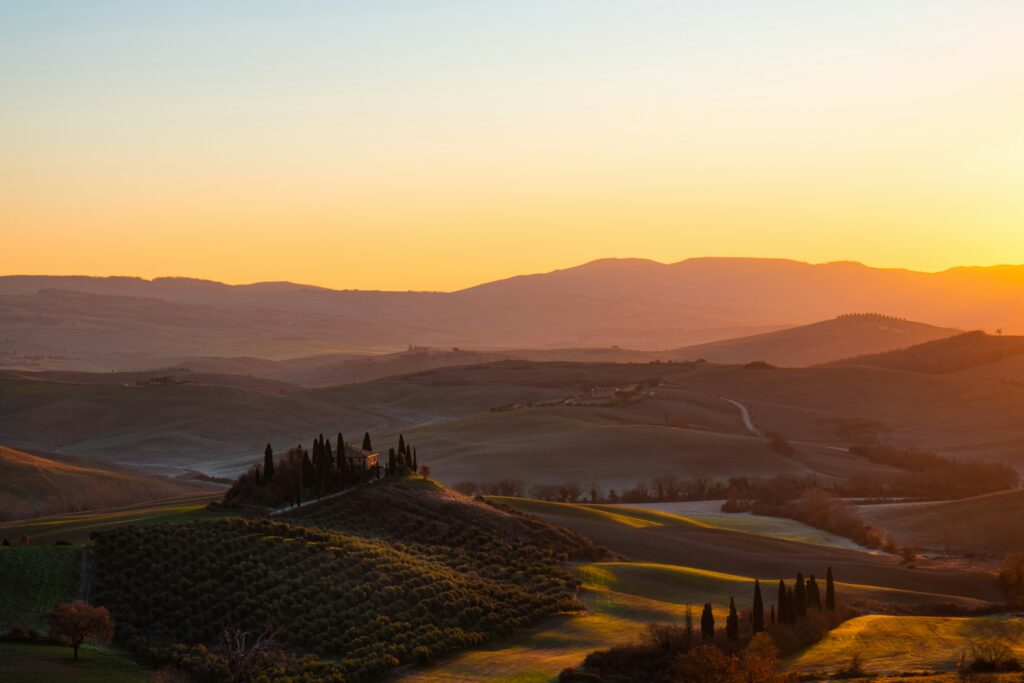 Panoramic golden hour landscape of rolling hills in Tuscany, Italy, featuring a traditional stone villa surrounded by tall cypress trees on a hill crest, bathed in warm orange and golden sunlight.