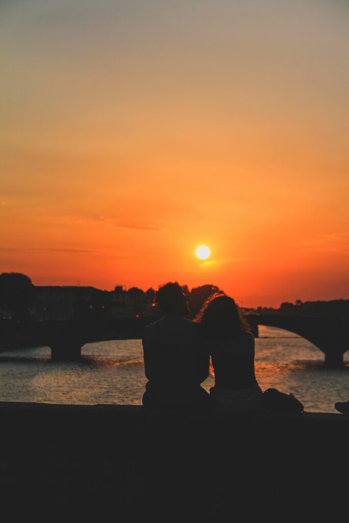 A romantic vertical shot of a man and a woman in silhouette, sitting by the stone edge of the Arno River in Florence, Italy. The sun is setting directly between them, creating a vibrant orange and golden sky. In the background, the soft silhouettes of Florence's historic bridges stretch across the calm water at twilight.