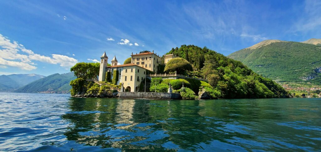 A wide landscape view from the water looking at a historic Italian villa with twin stone towers, manicured green terraces, and shaped trees on a lush peninsula over Lake Como, Italy. Deep blue water ripples in the foreground reflecting the clear sky with light wispy clouds, capturing the ultra-exclusive luxury travel experience.