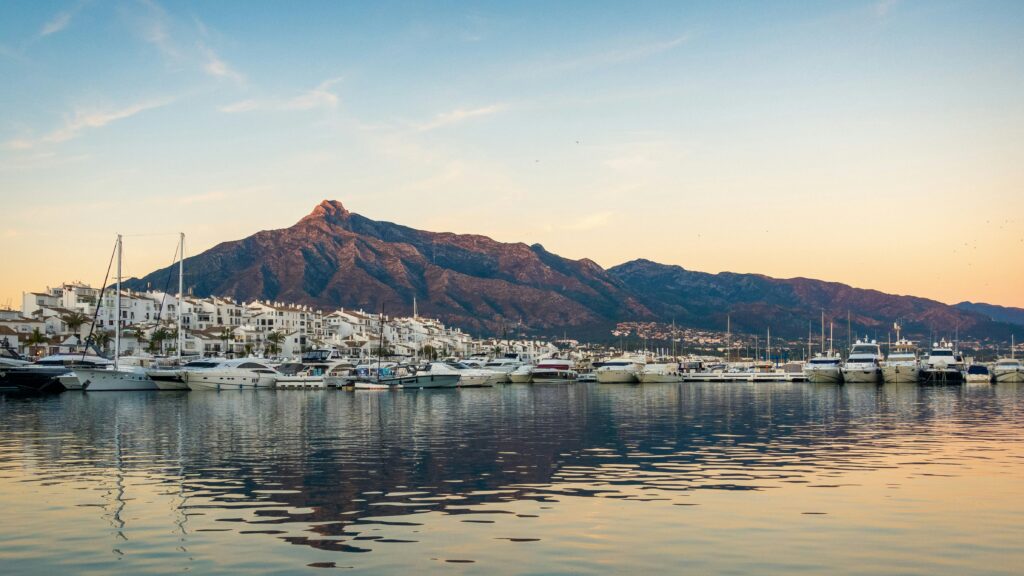 Panoramic sunset view of the luxury marina in Puerto Banús, Marbella, featuring white yachts docked along the harbor with whitewashed Andalusian buildings and the majestic La Concha mountain in the background.