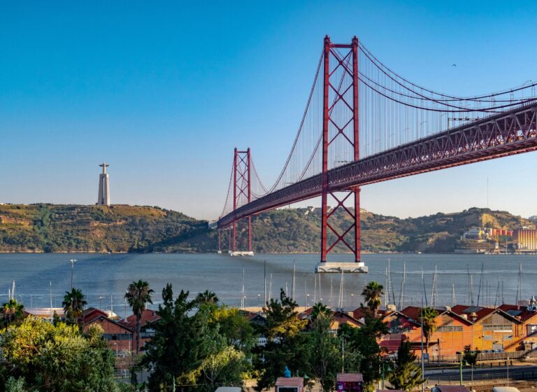A wide landscape view of the red 25 de Abril suspension bridge stretching across the Tagus River in Lisbon, Portugal, on a clear, sunny day. The Cristo Rei monument stands on the far hillside across the water, with a marina and green trees in the foreground.