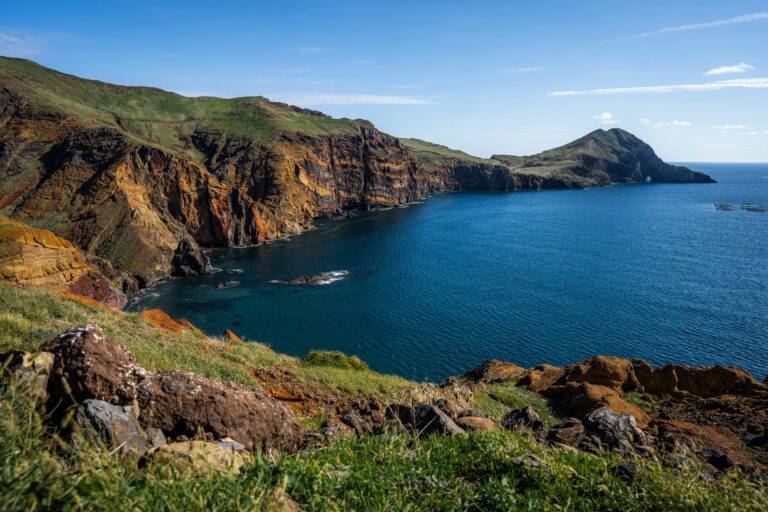 A landscape photograph of the rugged coastline of Madeira, Portugal, showcasing dramatic multicolored volcanic cliffs meeting the deep blue ocean under a clear sky, with grassy foreground slopes and rocky terrain.