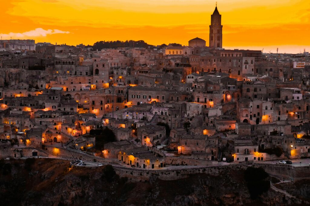 An aerial landscape photograph showing the ancient city of Matera, Italy, with its cave dwellings (Sassi) clinging to a steep hillside. The sky above is a vibrant, fiery orange at sunset, while the stone buildings below are bathed in shadow and illuminated by countless warm, glowing lights from windows and street lamps, creating a dramatic and historic atmosphere.