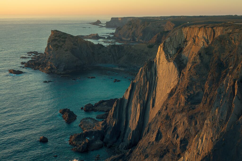Dramatic vertical cliffs of the Alentejo coast in Portugal during sunset, showing rock formations jutting into the turquoise Atlantic Ocean under a soft gradient sky.