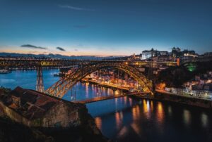 A dusk landscape photograph of the iconic double-deck Dom Luís I Bridge spanning the Douro River in Porto, Portugal, illuminated by warm city lights reflecting on the water with the historic city skyline in the background.